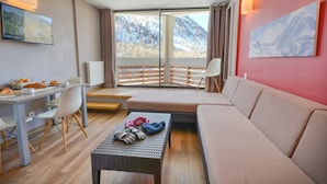 Une chambre d’hôtel moderne avec un grand lit, un canapé, une table à manger avec des chaises, une petite table basse et une vue sur une montagne enneigée par de grandes fenêtres.