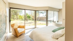 Une chambre avec un grand lit, un fauteuil en osier et une vue sur un jardin, visible à travers des portes-fenêtres.