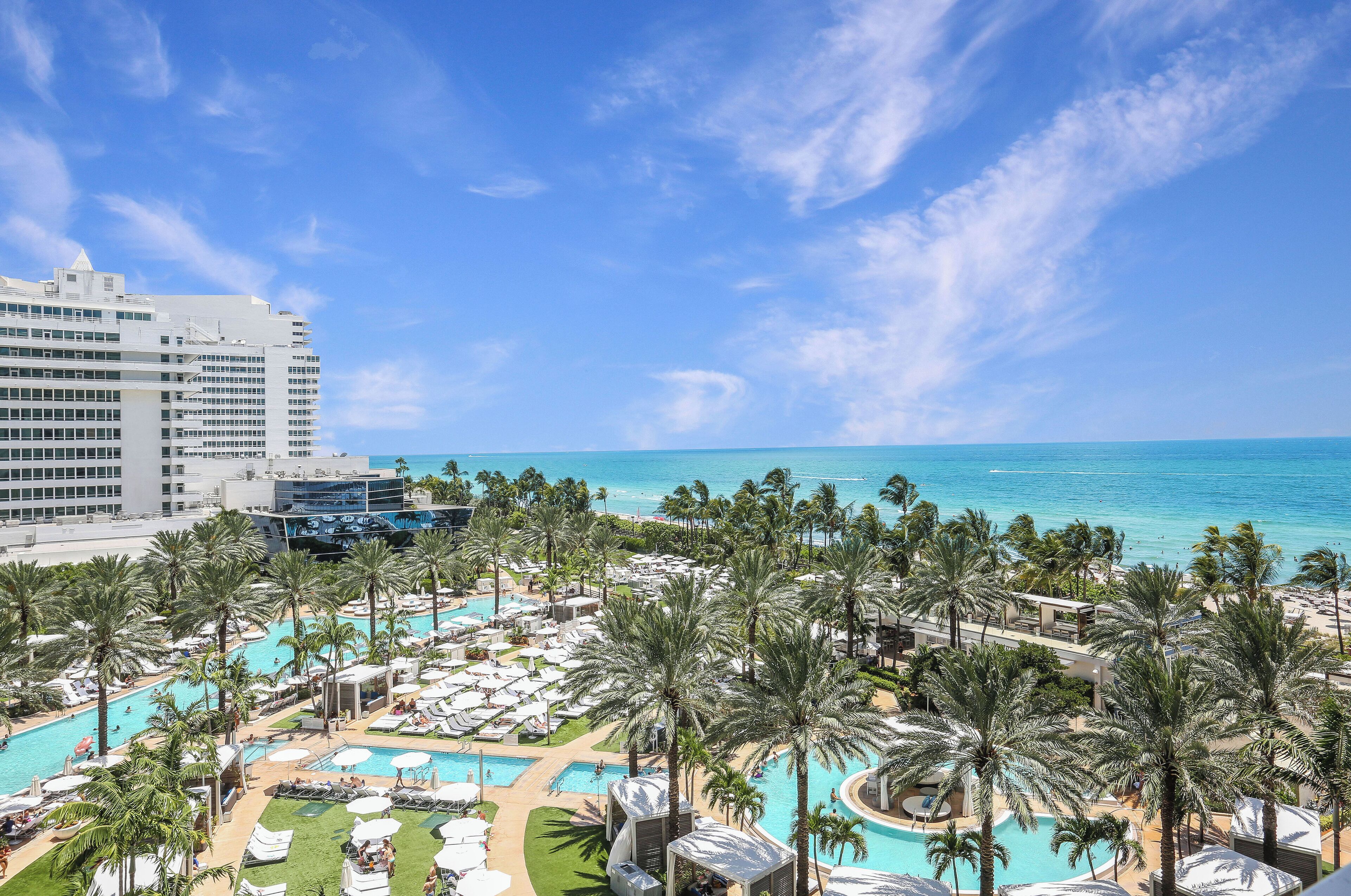 Un resort con un amplio área de piscina rodeada de palmeras, una playa y un cielo azul despejado.