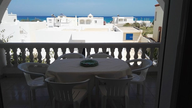 En balkon med et bord og stole, der giver udsigt over kysten med bygninger og havet.