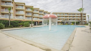 En swimmingpool med en svampeformet fontæne, omgivet af et flere etagers hotel med balkoner og palmer.