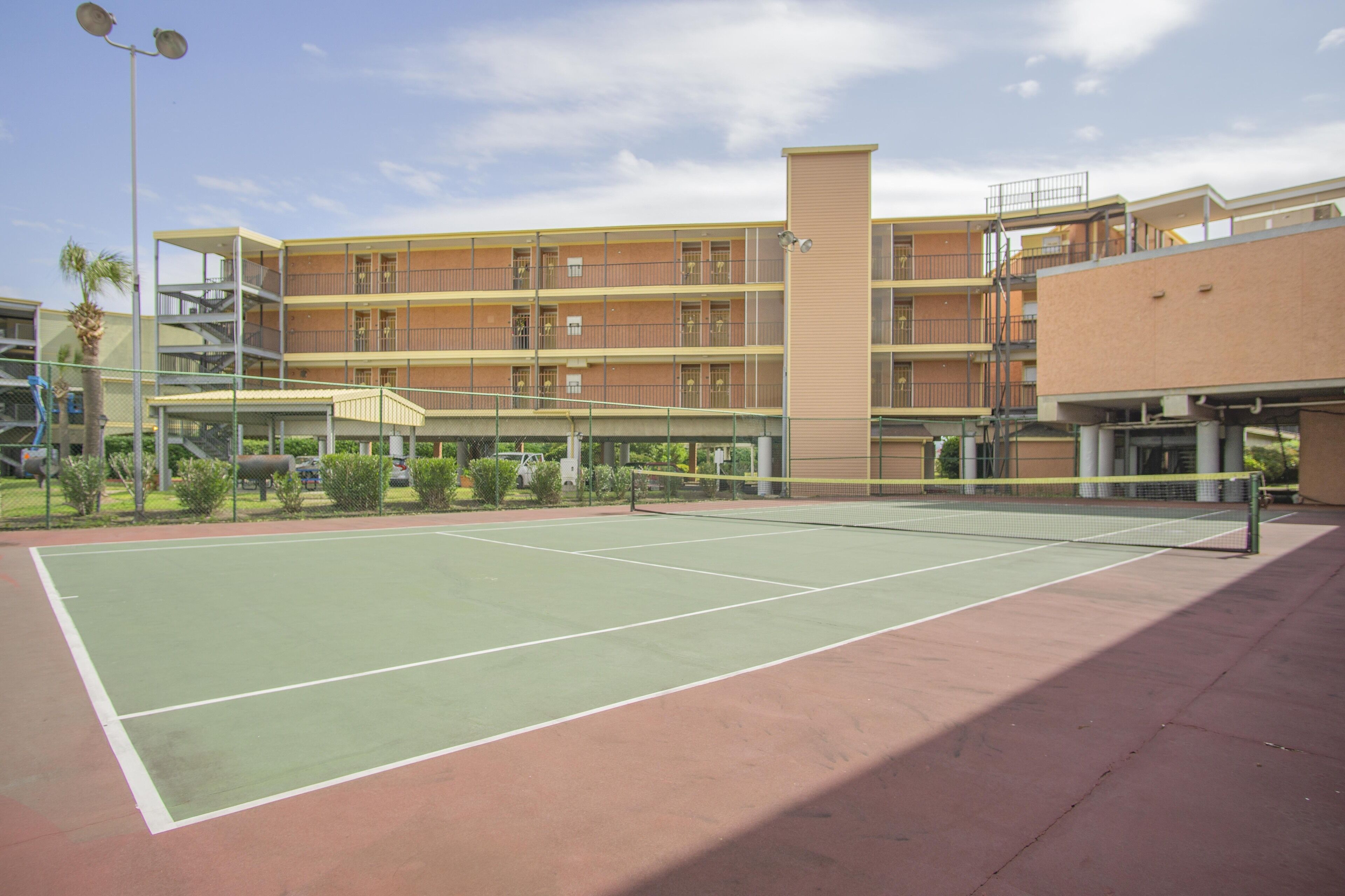 A tennis court with a net, surrounded by a building with multiple balconies and a covered walkway.