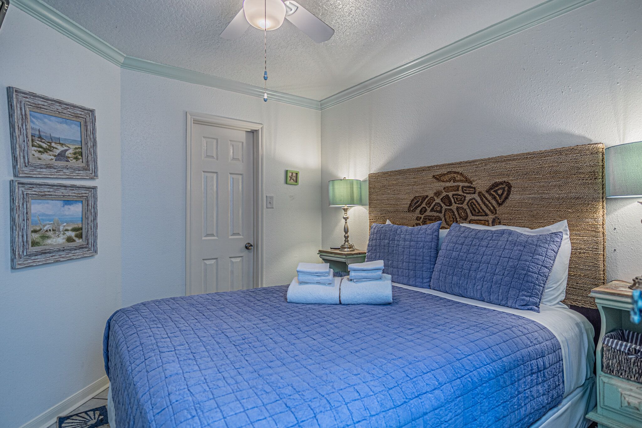 A bedroom with a large bed, a ceiling fan, framed pictures on the wall, and a nightstand with a lamp.