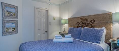 A bedroom with a large bed, a ceiling fan, framed pictures on the wall, and a nightstand with a lamp.