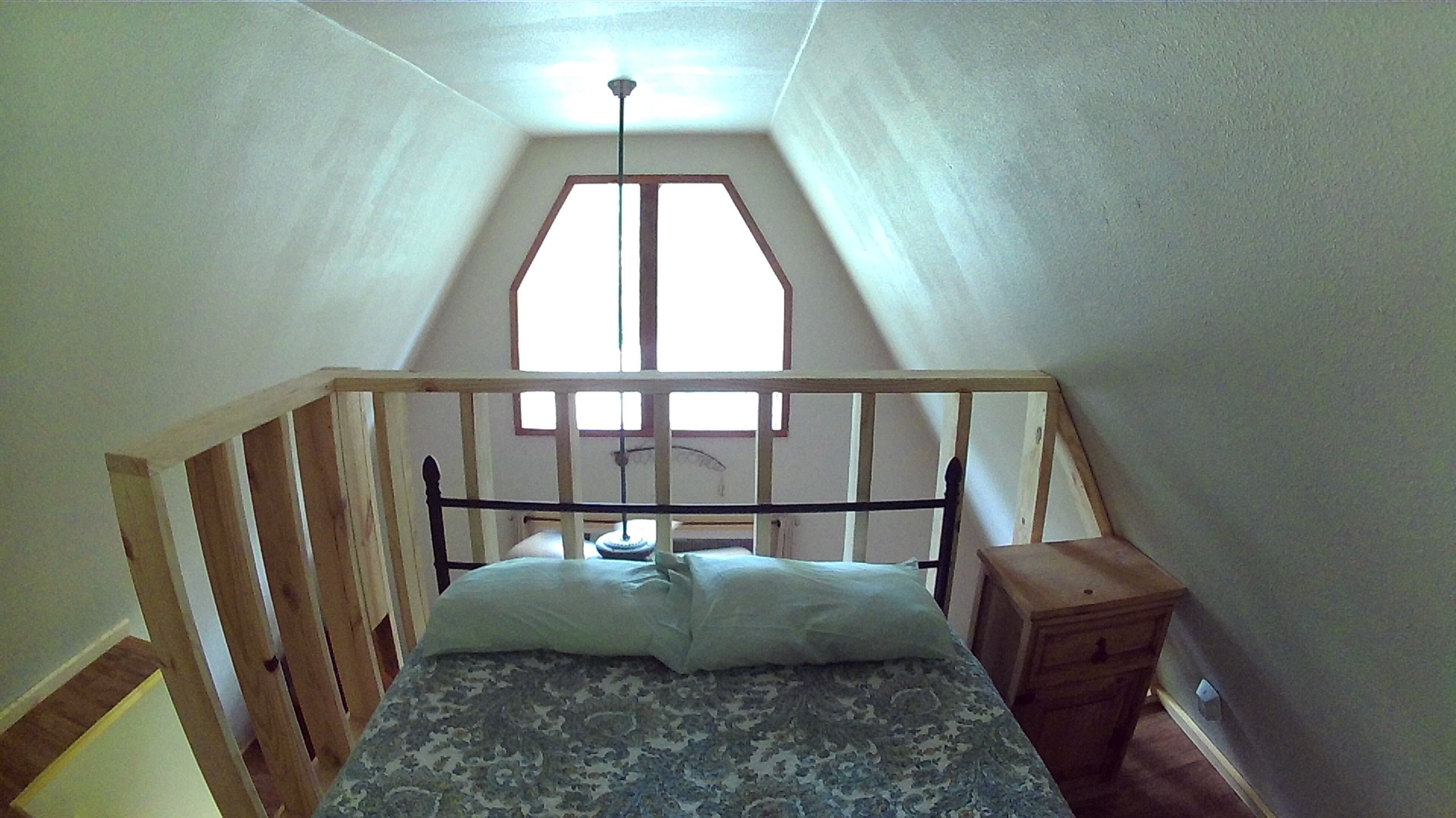 Un dormitorio bajo los tejados, con techo inclinado, estructura de cama de madera, una mesita de noche y una ventana con vista al cielo.
