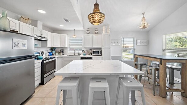 A modern kitchen with white cabinetry, a central island with stools, stainless steel appliances, and pendant lighting. - Beach Cottage with Soaking Pool & Outdoor Oasis (Galveston)
