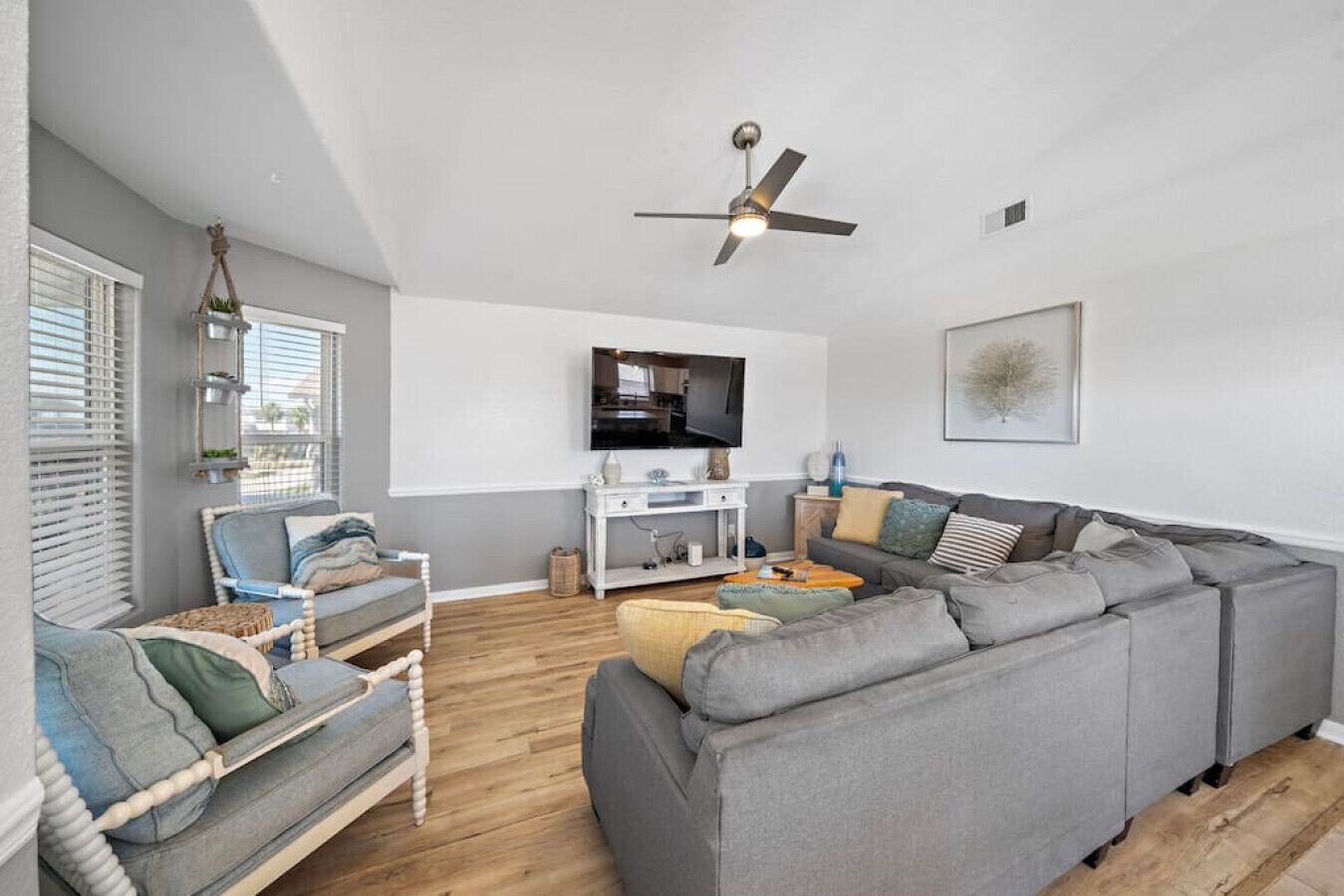 Een moderne woonkamer met een grijze hoekbank, een witte fauteuil en een houten salontafel. Er is een plafondventilator, een flatscreen televisie aan de muur en een raam met jaloezieën.