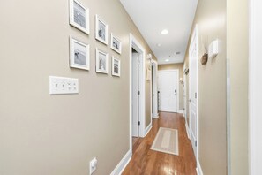 Un pasillo con paredes color beige, detalles blancos y suelo de madera. Se observan cuadros enmarcados en la pared y una puerta al final.