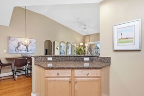A kitchen with granite countertops, wooden cabinets, a dining area with chairs, a hanging light fixture, and a framed landscape painting on the wall.