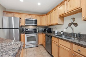 A kitchen with wooden cabinets, stainless steel appliances including a refrigerator, microwave, oven, and dishwasher, a granite countertop, and a sink with a modern faucet. - Greyt Escape: Lovely, Ocean View Condo Across from OKI Pier with Community Pool, (Oak Island)