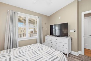 A bedroom with a bed, a dresser, a television, and a window with curtains. - Greyt Escape: Lovely, Ocean View Condo Across from OKI Pier with Community Pool, (Oak Island)