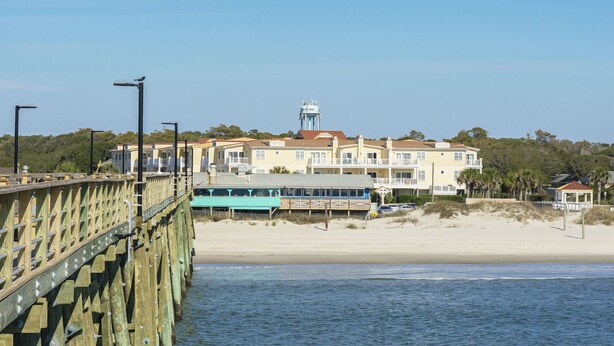 Un hotel costero con varios balcones, vista a la playa y un muelle que se adentra en el agua.