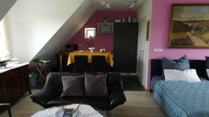 Una habitación con un sofá de cuero negro, una cama con colcha azul, una mesa de comedor con mantel amarillo y un ventanal pequeño.