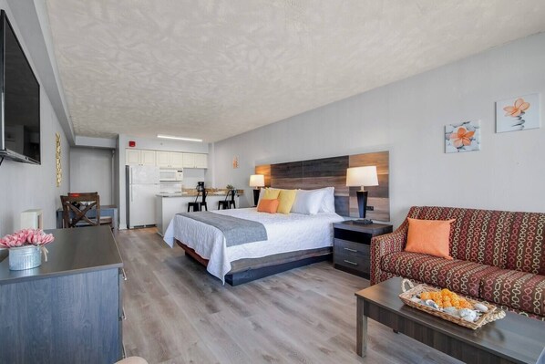 A modern hotel room with a bed, a sofa, a dining table, and a kitchen area.