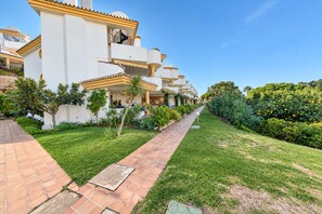 Una hilera de edificios blancos con balcones, un sendero pavimentado y abundante vegetación.