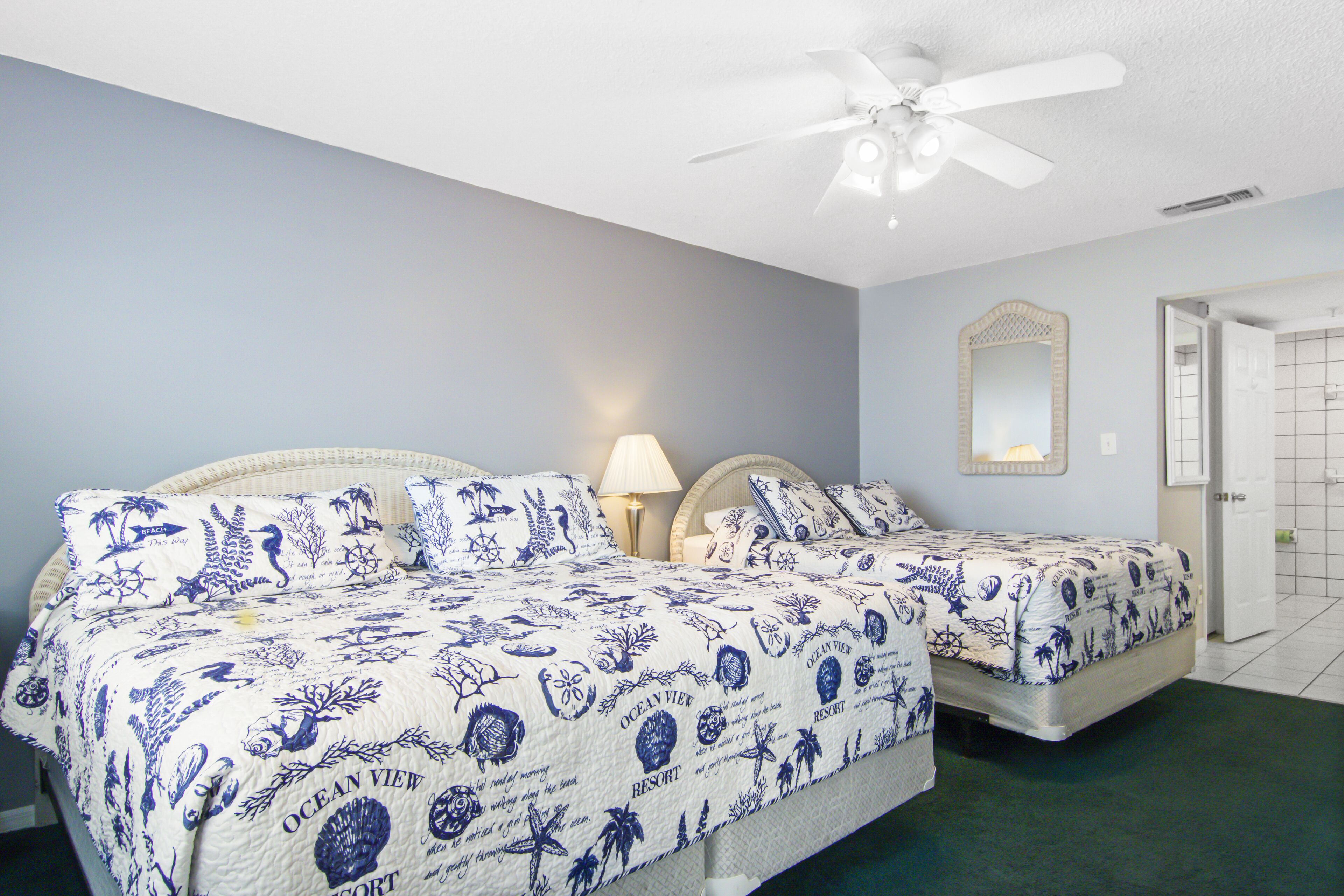 A bedroom with two beds, a ceiling fan, a nightstand with a lamp, and a mirror on the wall.