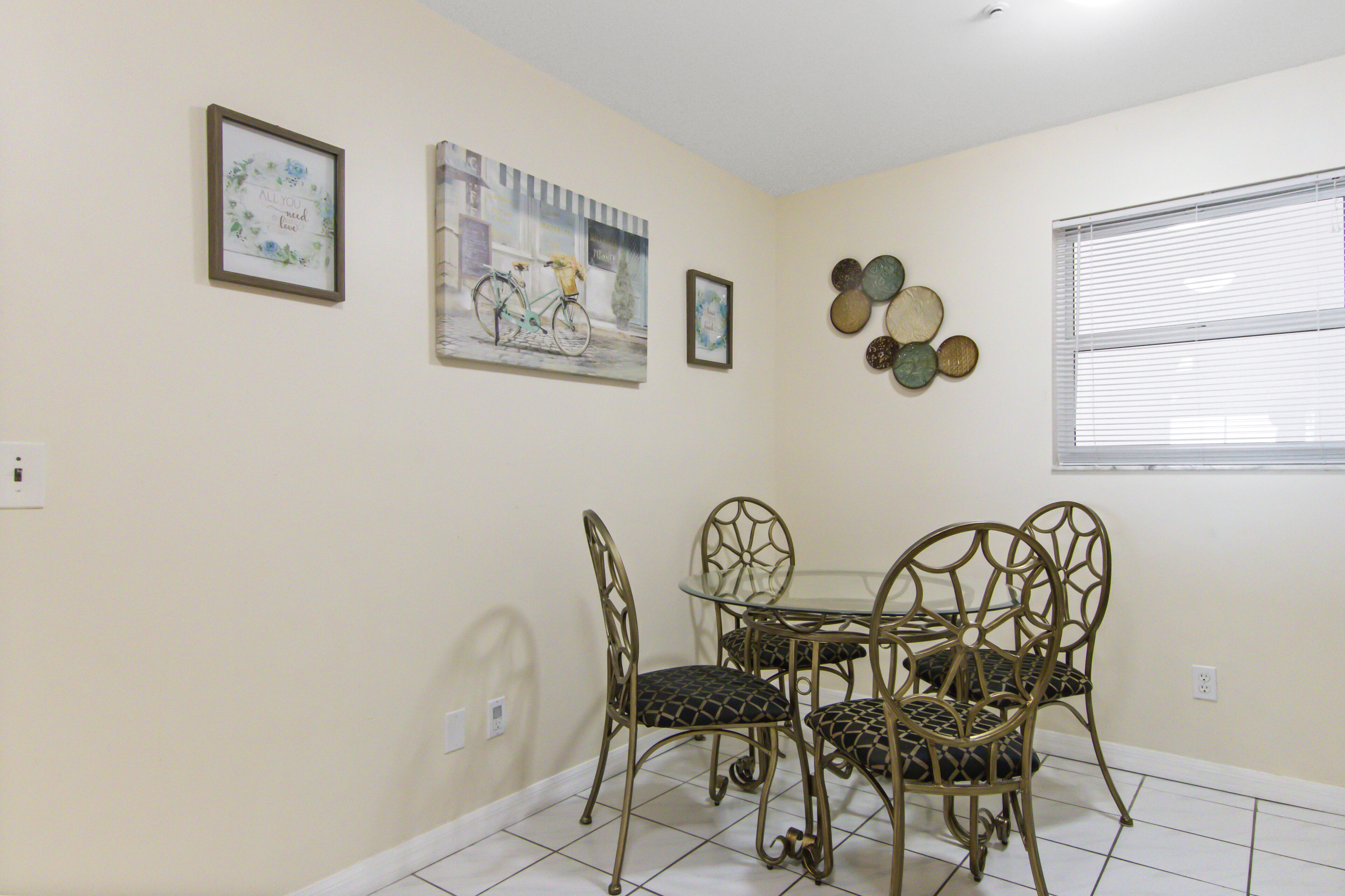Un comedor con una mesa de vidrio y cuatro sillas, un cuadro en la pared, y una ventana con persianas.