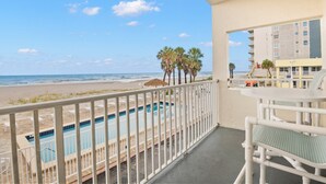 A balcony with a view of a swimming pool and beach, a table with chairs, and a building in the background.