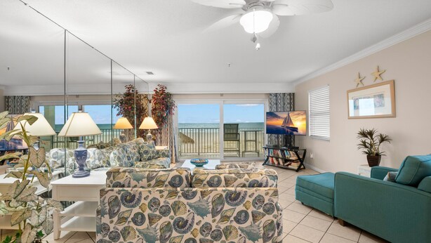 A spacious living room with a large sofa, a smaller sofa, a dining table with chairs, and a ceiling fan. There is a large window with a view of the ocean.