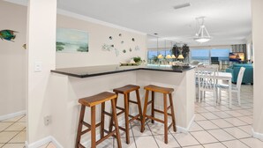 A kitchen with a bar area, white walls, and a tiled floor. There are three wooden stools, a white dining table with chairs, and a view of the ocean through large windows.