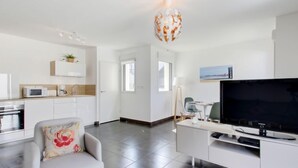 A modern living room with a flat-screen TV, a dining area with a table and chairs, a kitchen with white cabinets and appliances, and a decorative hanging light fixture.