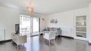 A modern living room with a grey sofa, white armchairs, a coffee table, and a TV on a white cabinet. There is a glass door leading to an outdoor area.