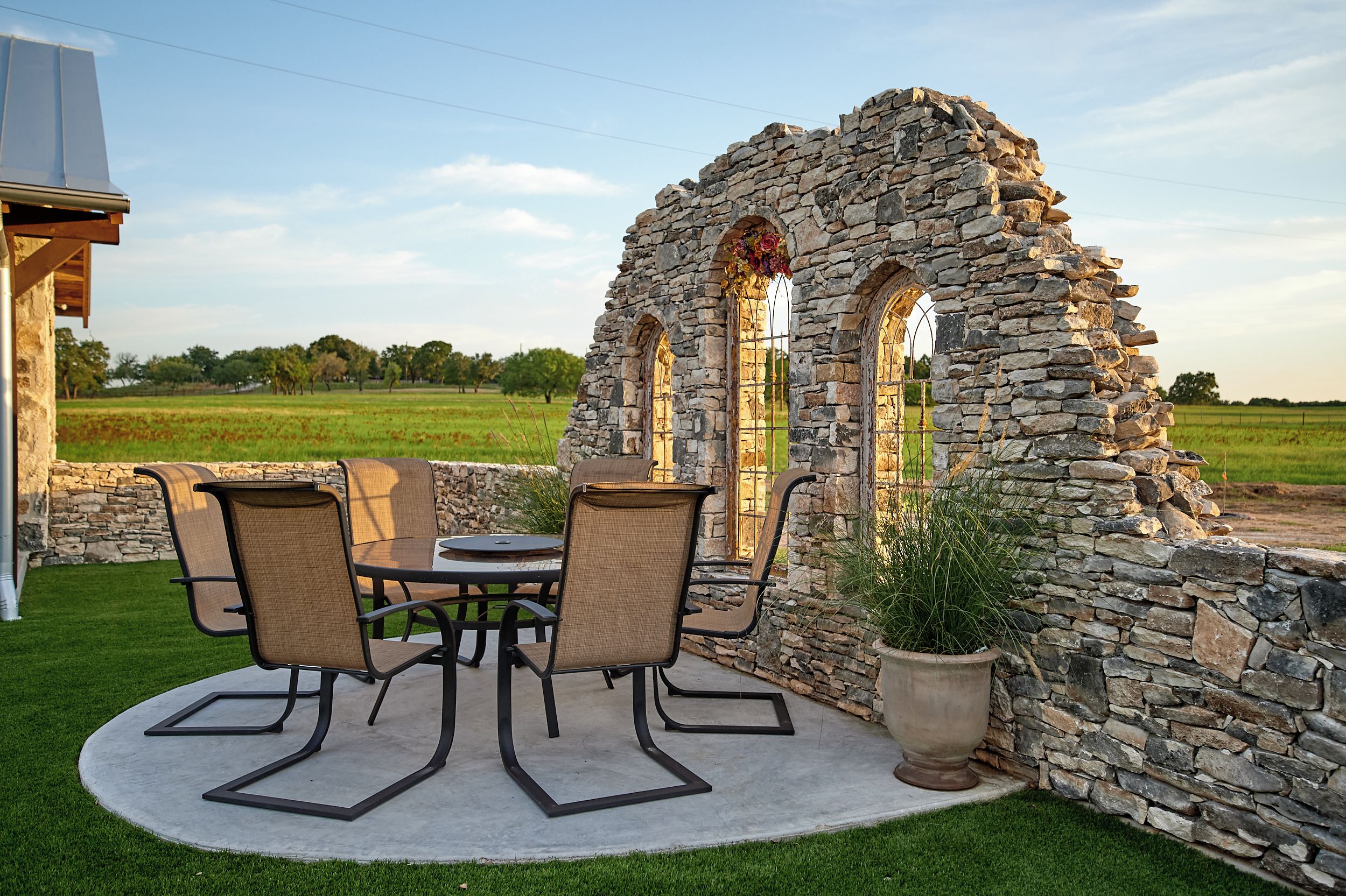 Une structure en pierre dotée de fenêtres en arc de cercle, une table ronde avec des chaises et une plante en pot.