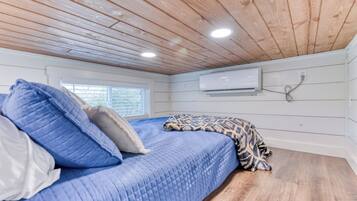 Un dormitorio con techo de madera, cama con ropa de cama azul, manta estampada y un equipo de aire acondicionado en la pared.