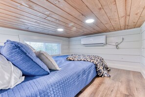 Un dormitorio con techo de madera, cama con ropa de cama azul, manta estampada y un equipo de aire acondicionado en la pared.