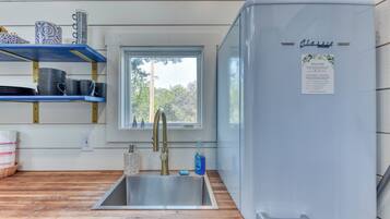 Una cocina con un lavamanos de acero inoxidable, un refrigerador azul y estantes con platos y tazas.