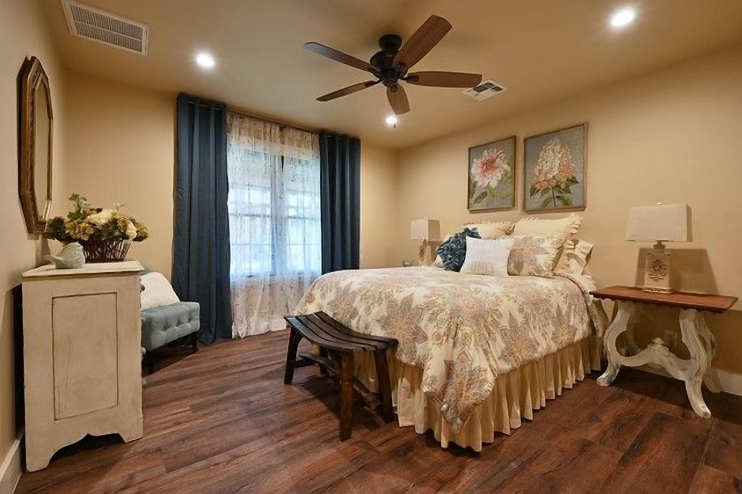 A bedroom with a bed, a chair, a nightstand, a ceiling fan, and a window with curtains.
