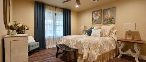 A bedroom with a bed, a chair, a nightstand, a ceiling fan, and a window with curtains.