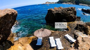 Un paysage côtier avec une eau bleue et limpide, un terrain rocheux et des chaises longues placées sous un parasol en chaume.