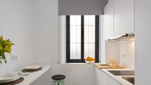 Une petite cuisine avec une fenêtre, des placards blancs, deux tabourets et un saladier rempli d’oranges posé sur le comptoir.
