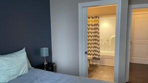 A bedroom with a bed, a nightstand, a lamp, and a bathroom visible through an open door.