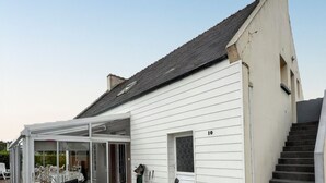 A white building with a covered patio area featuring outdoor furniture and a table. - Homerez - House in Cléder. (Cléder)