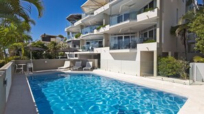 A modern hotel with a swimming pool, lounge chairs, and a clear blue sky.