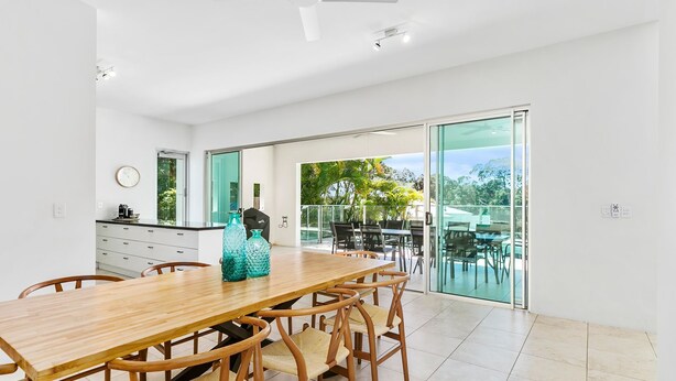A modern dining area with a wooden table and chairs, a kitchen counter with appliances, and a sliding glass door leading to an outdoor patio with a view of greenery.
