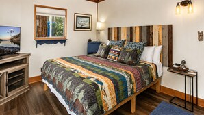 A bedroom with a wooden headboard, a large bed with patterned bedding, a television on a wooden stand, and a framed picture on the wall.