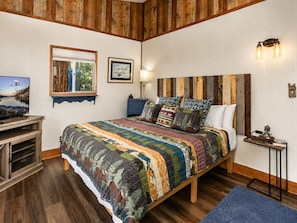 Ein Schlafzimmer mit einem Holz-Kopfteil, einem großen Bett mit gemusterten Betttextilien, einem Fernseher auf einem Holzschrank und einem gerahmten Bild an der Wand.
