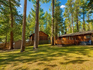 A cabin surrounded by tall trees, with a wooden deck and a small shed nearby.