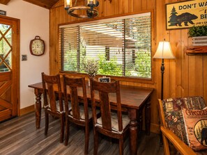 A wooden interior with a dining table, chairs, a clock, a window with blinds, a lamp, and a sign.