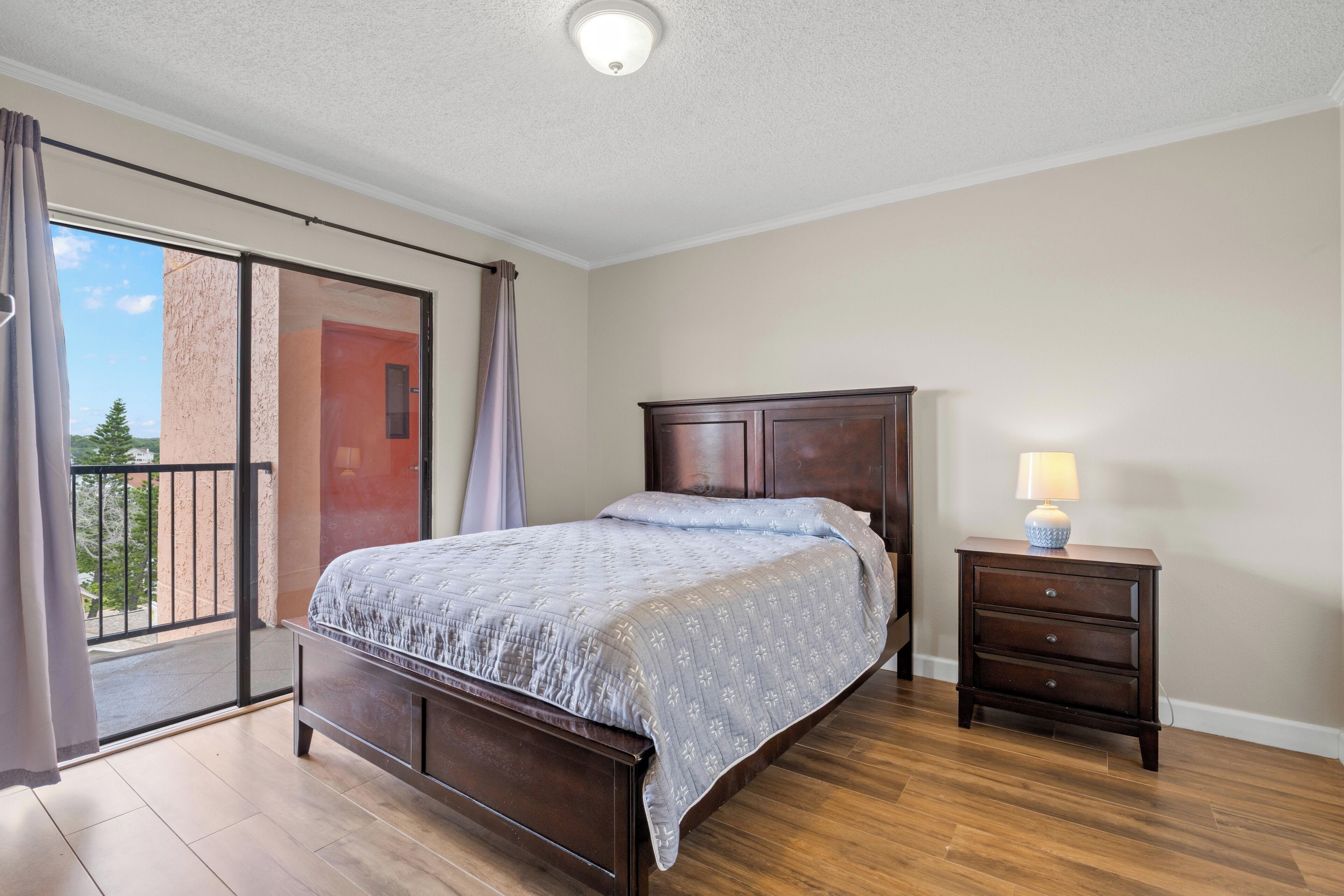A bedroom with a bed, a nightstand, a lamp, a sliding glass door, and a view of greenery outside.