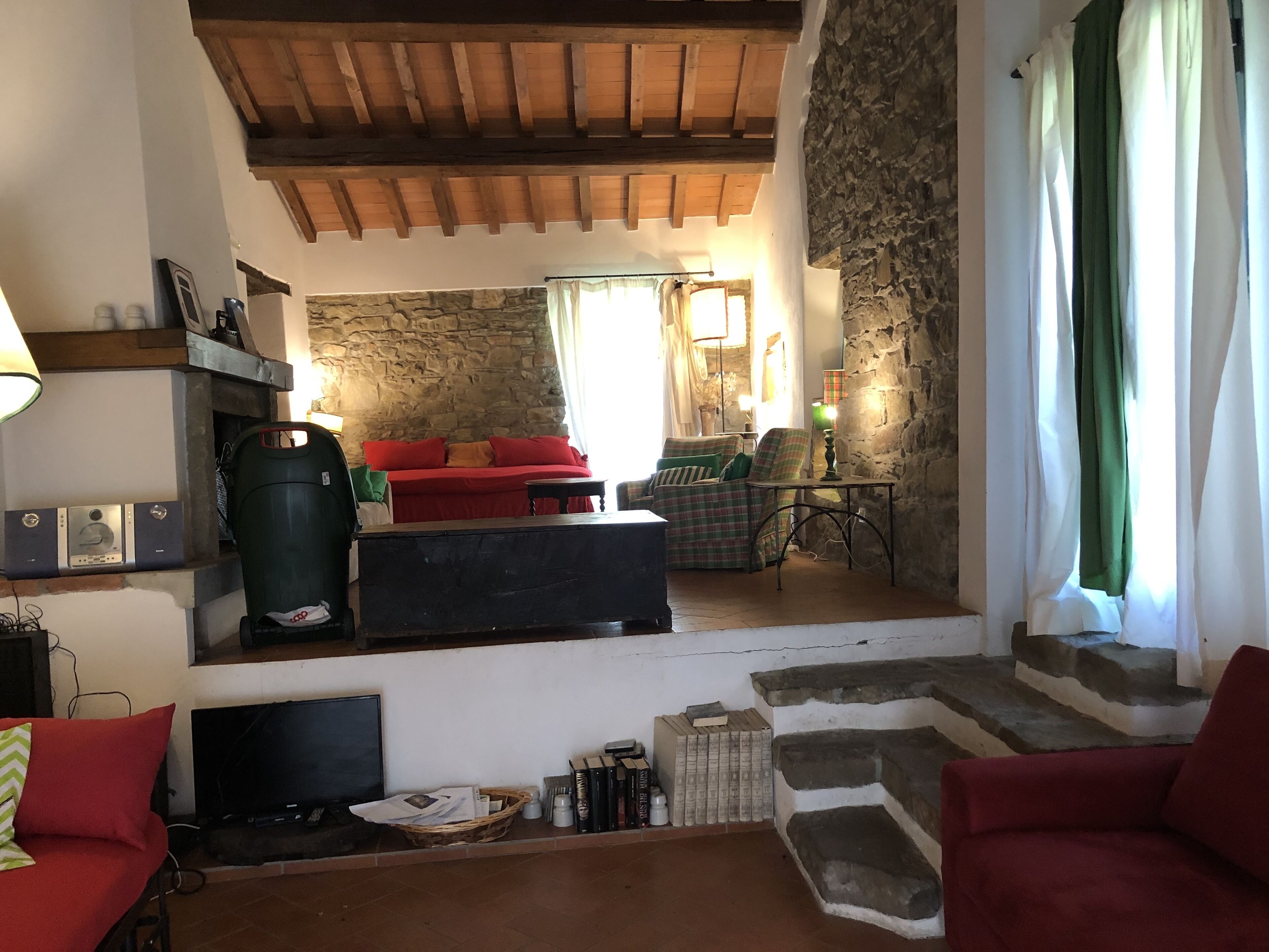 A cozy living room with a stone wall, wooden ceiling beams, a red sofa, a green armchair, and a television.