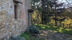 Ein Steinhaus mit rustikalem Äußeren, einer hölzernen Bank und einem Kiesweg, der zu einem von Bäumen gesäumten, eingezäunten Bereich führt.