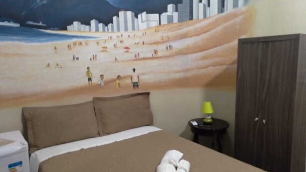 Una habitación de hotel con una cama, un mural en la pared que representa una playa, una mesita con una lámpara y un armario.