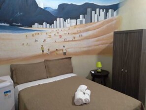 A hotel room with a bed, a wall mural of a beach scene, a small table with a lamp, and a wardrobe. - Hotel Nacional (Sobral)