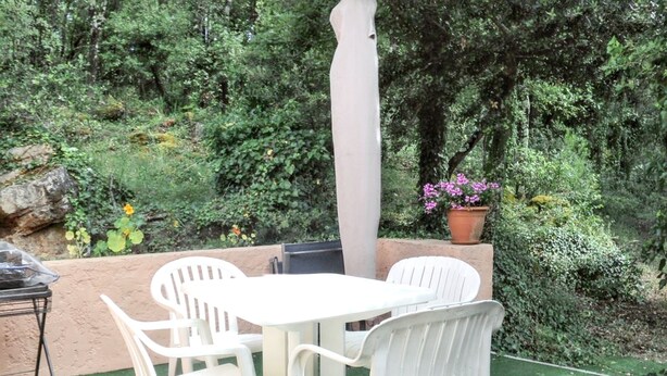 Un patio al aire libre con una mesa redonda y cuatro sillas blancas, rodeado de follaje y una maceta con flores rosadas.