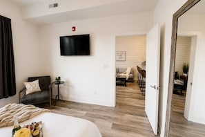 Habitación de hotel con cama, televisor montado en la pared, una mesita con una planta y un acceso a un comedor.