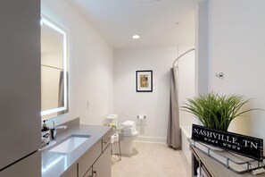 Un baño moderno con un espejo grande, lavamanos dobles, inodoro y ducha. Hay un cuadro en la pared y un letrero que indica la ubicación como Nashville, TN.